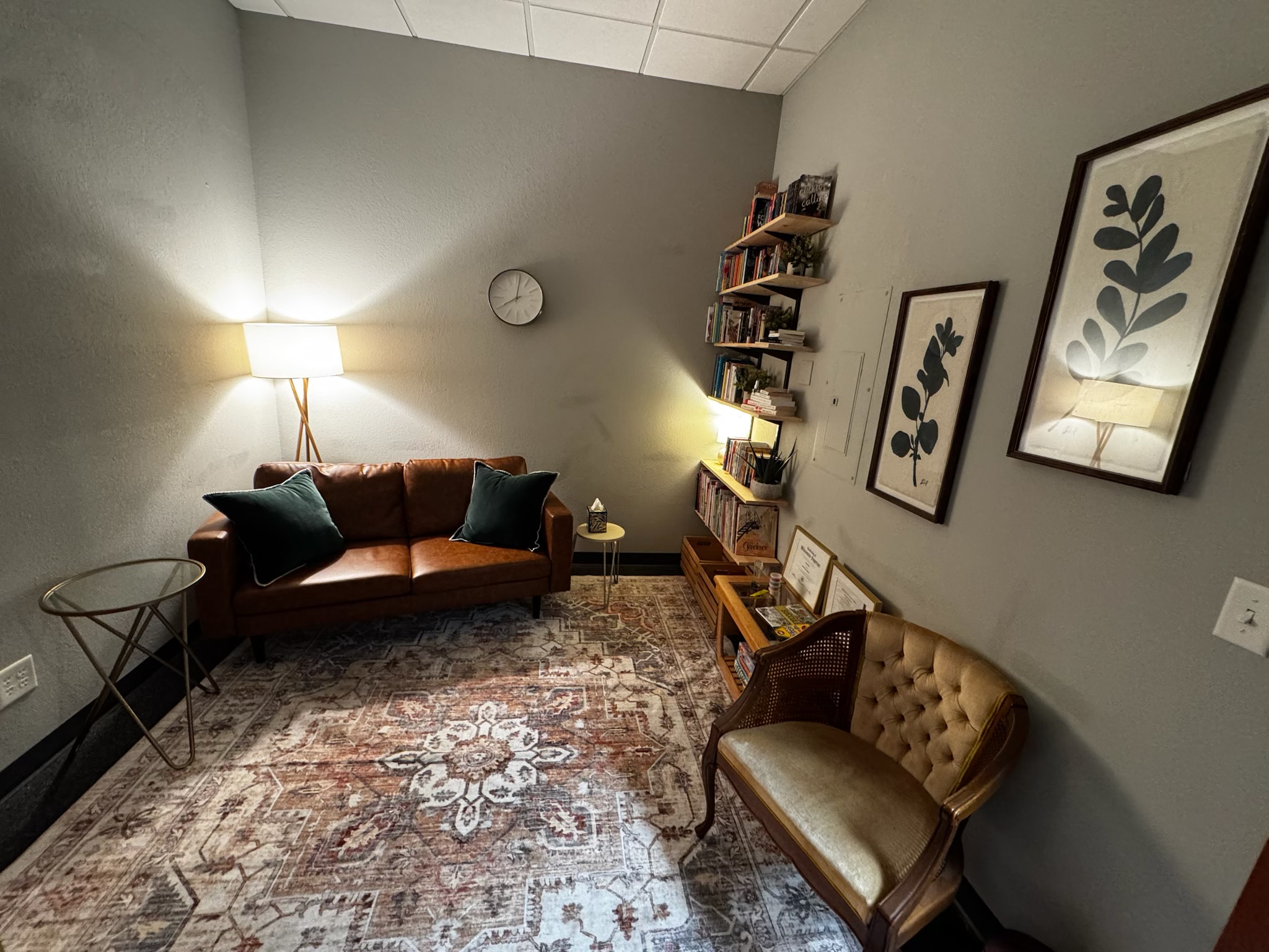 The therapy room at the Superior office, with a leather couch, floor lamp, area rug, framed botanical prints, and a vintage upholstered chair.